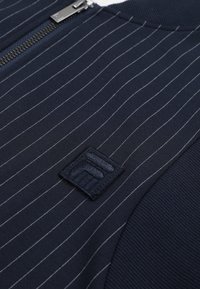 Tissu bleu marine à fines rayures avec une texture côtelée sur les bords, doté d'une fermeture éclair en métal et d'un petit patch brodé avec un motif géométrique.