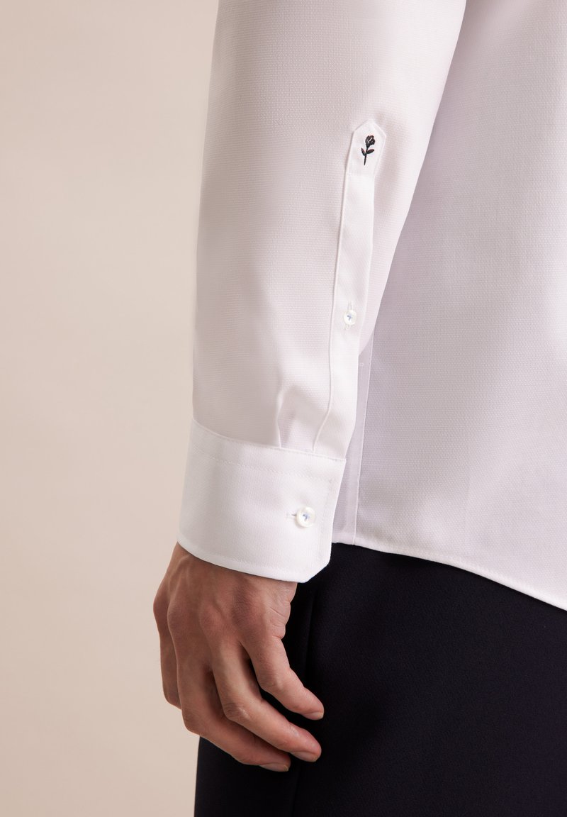 Witte blouse met een geweven stof, knoopmanchetten en een kleine zwarte rozenborduursel nabij de manchette.
