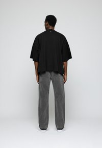 Hombre de espaldas vistiendo una camiseta negra holgada, vaqueros grises y zapatillas blancas frente a un fondo liso gris claro.