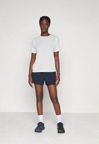 Valge lühikeste varrukatega sportlik t-särk, kombineeritud tume mereväe siniste lühikestega. Modell kannab musti kingi ja valgeid sokke, seistes lihtsa tausta ees.