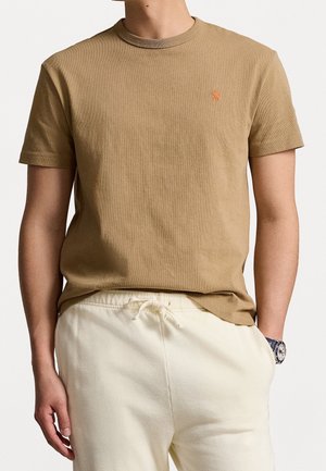 Tan bomulds T-shirt med korte ærmer, ribbet rund halsudskæring og lille orange logo på brystet. Bæret sammen med hvide sweatpants med snøring.