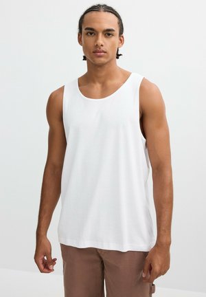 Joven con cabello trenzado que lleva una camiseta sin mangas blanca lisa y pantalones marrón claro, de pie frente a un fondo claro y simple.