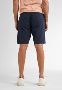 Pantalones cortos azul marino fabricados en un material ligero, que cuentan con dos bolsillos traseros con botones y un corte relajado, combinados con zapatillas beige.