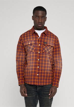 Giovane uomo con camicia a quadri rossa e arancione abbottonata sopra una t-shirt bianca, abbinata a jeans scuri lavati, in piedi davanti a uno sfondo semplice.