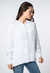White button-up shirt made of textured fabric, featuring a collar, long sleeves, and a relaxed fit with a slightly curved hem.