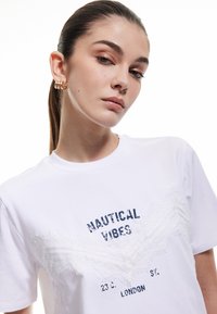 Hvid bomulds t-shirt med "NAUTICAL VIBES" i marineblå tryk og struktureret hvid blonde detaljer forneden. Rund hals og korte ærmer.