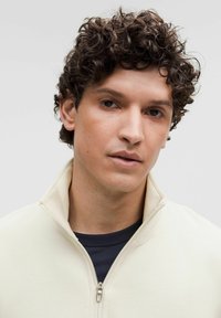 Felpa zip-up beige chiaro con colletto alto, tessuto strutturato e camicia blu scuro sotto. Il modello ha capelli ricci e scuri.