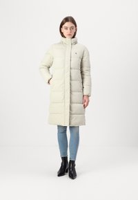 Light gray puffer coat with vertical quilted design, high collar, and logo detail, worn with light blue jeans and black ankle boots.
