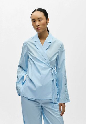 Aziatische vrouw in een lichtblauwe overslagblouse met geborduurde lange mouwen en bijpassende broek, staand tegen een witte achtergrond.