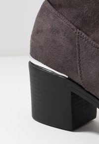 Botín gris de ante con un tacón grueso texturizado en negro y un acento metálico plateado alrededor de la base del tacón.