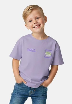 Glimlachende blonde jongen met een lavendelkleurig T-shirt met tekst, staand met handen in de zakken en een blauwe spijkerbroek tegen een effen lichte achtergrond.