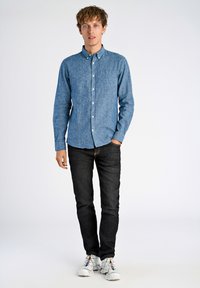 Lichtblauw overhemd met textuur, borstzak, gecombineerd met donkere spijkerbroeken en witte sneakers met kleurrijke accenten.