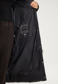 Interno di un giubbotto imbottito nero, realizzato in poliestere riciclato al 100%. Presenta un'etichetta con simboli, chiusura con zip e bottoni a pressione lungo l'orlo.