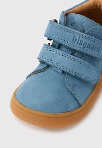 Chaussure en daim bleu avec semelle en caoutchouc marron, dotée de deux brides réglables à scratch et de coutures contrastées. Nom de la marque embossé sur la bride.