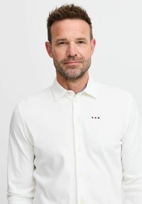 Cămașă albă cu mâneci lungi, tip cămașă, fabricată dintr-un material moale, având un logo brodat subtil cu accente roșii și albastre pe piept.