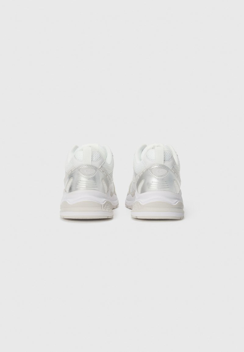 White sneakers with a glossy silver accent feature a mesh upper, textured detailing, and a flexible white sole with a subtle design.