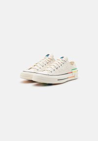 Płótno sneakersy w odcieniu off-white z drobnymi kropkami, kontrastującą czarną podeszwą oraz kolorowymi paskami po bokach. Posiada białe sznurowadła i metalowe oczka.