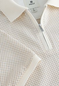 Polo à carreaux beige et blanc avec une demi-fermeture éclair, un col texturé et un tissu doux. L'étiquette indique "NEXT" et la taille "M".