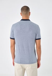 Man som står med ryggen mot kameran, iklädd en ljusblå kortärmad polo med mörk krage och ärmslut, i kombination med ljusbeiga byxor.