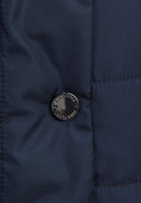 Navyblå pufferjacka i tyg med en slät textur. Har en rund, glänsande svart knapp präglad med "PINA PLAGE" nära sömmen.