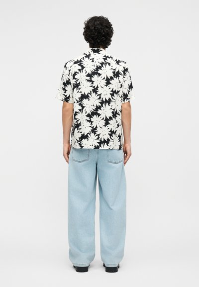 Homme aux cheveux bouclés portant une chemise à manches courtes noire et blanche à motifs floraux et un jean large bleu clair, vu de dos.