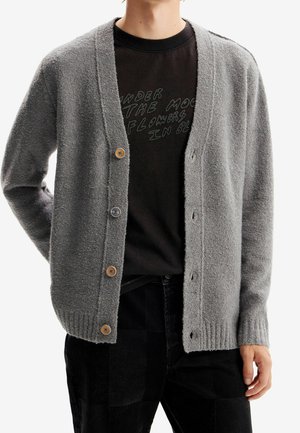 Personne portant un cardigan gris boutonné sur un T-shirt noir avec un texte partiellement visible, associé à un pantalon noir.