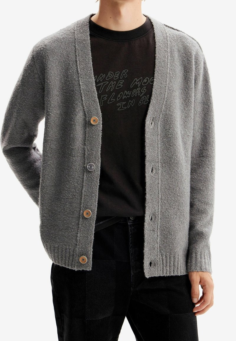 Personne portant un cardigan gris boutonné sur un T-shirt noir avec un texte partiellement visible, associé à un pantalon noir.