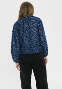 Vrouw met lang haar draagt een gestructureerde donkerblauwe panterprint top en zwarte broek, gezien vanuit de rug tegen een effen witte achtergrond.