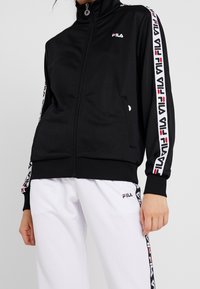 Veste noire zippée en tissu lisse avec col montant. Présente des bandes de marque blanches et rouges le long des manches. Poches latérales incluses.