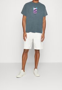 Baumwoll-T-Shirt in Dunkelblau mit lila Grafikdruck. Kombiniert mit weißen Shorts und hellen Sneakers, die Schnürsenkel und ein minimalistisches Design aufweisen.