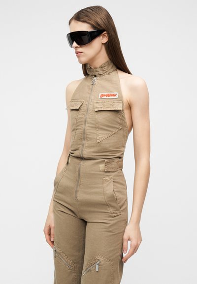 Femme portant une combinaison beige sans manches avec une fermeture éclair à l'avant et des poches, assortie de grandes lunettes de soleil noires futuristes.