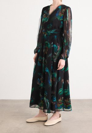 Lang sort kjole med blå og grønne fjerprint, gennemsigtige lange ærmer, tætsiddende talje, båret med beige flade sko på neutral gulv.