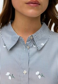 Camicia a righe con colletto morbido, colori chiari con accenti floreali verde e bianco, e tessuto texturizzato in vista ravvicinata.