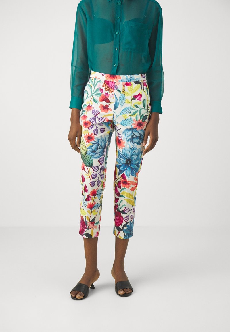 Personne portant un pantalon blanc court à imprimé floral coloré, une chemise longue turquoise transparente boutonnée et des talons ouverts noirs.