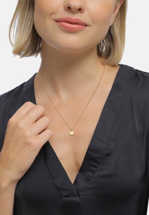 Goldenes Anhänger-Halsband mit einem runden Charm an einer zarten Kette, getragen über einem schwarzen V-Ausschnitt-Oberteil. Glatte Textur, minimalistisches Design.