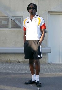 Sportaš u bijelom i crvenom Adidas dresu s žutim detaljima i crnim hlačicama, s naočalama za sunce i crnim tenisicama, stoji naslonjen na zid.