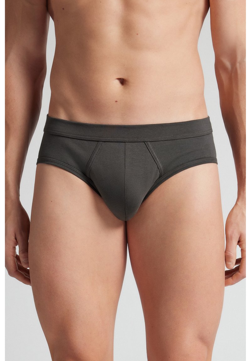 Torse masculin portant un slip gris foncé, debout avec les bras détendus le long du corps sur un fond uni.