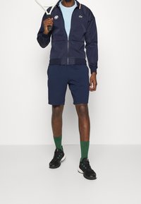 Man i marinblå sportjacka och shorts som håller en tennisracket över axeln, iklädd gröna strumpor och svarta sportskor.