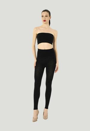 Vrouw staand, dragend een zwarte tube top, zwarte hoge taille leggings en beige hoge hakken tegen een effen lichte achtergrond.