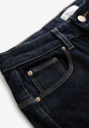 Vaqueros de denim oscuro con un pequeño bolsillo frontal, herrajes de cobre y costuras contrastantes a lo largo de las costuras. Textura suave con un leve brillo.