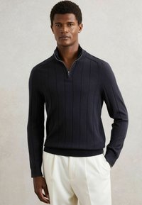 Ciemnoniebieski sweter z dzianiny z ćwierć zamkiem, z pionowymi paskami i ściągaczami przy rękawach. Miękka tekstura, dopasowany krój i kontrastowy brzeg.