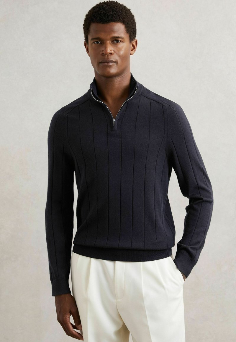 Ciemnoniebieski sweter z dzianiny z ćwierć zamkiem, z pionowymi paskami i ściągaczami przy rękawach. Miękka tekstura, dopasowany krój i kontrastowy brzeg.