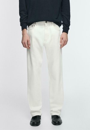 Osoba ubrana w luźne białe spodnie, czarne skórzane wsuwane buty oraz ciemną bluzkę z długim rękawem, stojąca na jednolitym tle.