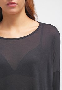 Woman wearing a sheer black top revealing a black bra underneath, showing neckline, shoulders, and upper chest.