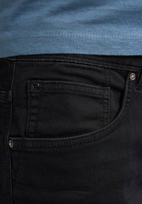 Schwarze Jeans aus Denim mit glatter Textur, die ein Detail mit Gesäßtasche und dezente Nähte aufweisen. Ein blauer T-Shirt ist leicht sichtbar darüber.