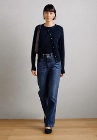 Model egy sötétkék gombos kardigánt, fekete felsőt, magas derekú kék farmert és fekete cipőt visel, semleges háttér előtt sétál.