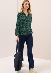 Groen gebloemd blouse met een V-hals en lange mouwen, gecombineerd met donkere jeans en een marineblauwe totebag. Model draagt lichte sneakers.
