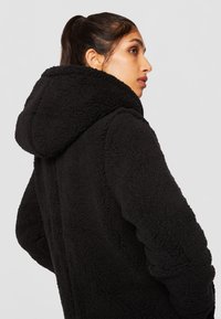 Veste en polaire noire avec capuche, au toucher doux et texturé, avec une coupe décontractée, vue de dos, mettant en avant le matériau et la forme.