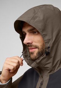 Bruine hoodie-jack met een gestructureerde afwerking en verstelbare trekkoorden. Heeft een contrasterend donker paneel aan de voorkant. Close-up weergave.