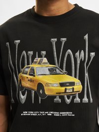 Melns T-krekls ar dzeltenā Ņujorkas taksometra grafiku un tekstu "Ņujorka". Taksimetrs ir attēlots detalizētā priekšējā skatā, un uz jumta ir ķekaru raksts.